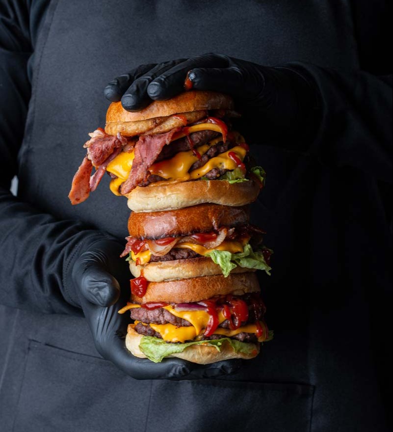 Stacked gourmet burger held by a chef wearing black gloves, showcasing layers of beef, bacon, cheese, and salad