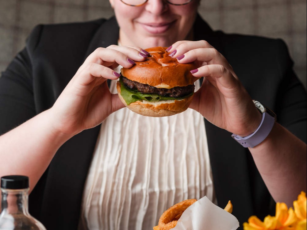 Person wearing glasses and a black blazer about to take a bite of a burger at a table with fries, drinks, and condiments.