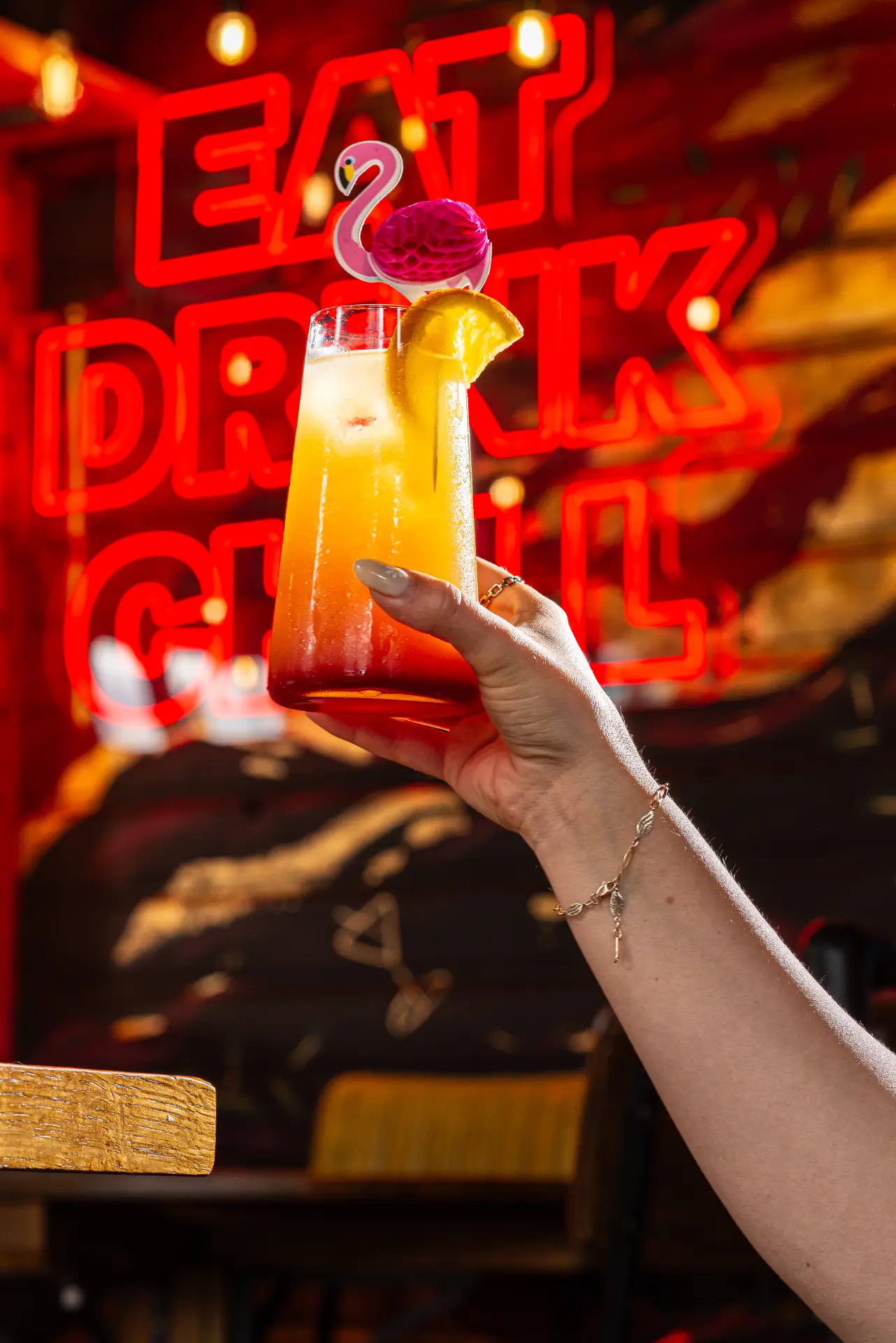 Hand holding a tall layered orange cocktail garnished with an orange slice and a pink flamingo topper in front of a red "Eat Drink Chill" neon sign at The Fat Lounge in Cambridge