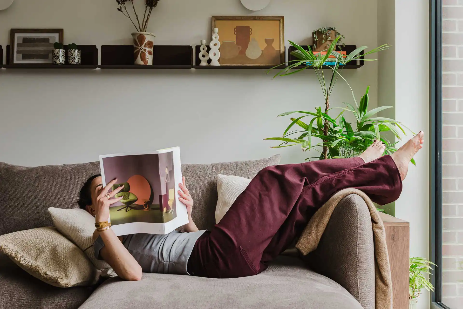 A person lying comfortably on a large grey sofa with bare feet propped up on the armrest, reading a magazine that partially covers their face in a bright living room with shelving and plants.