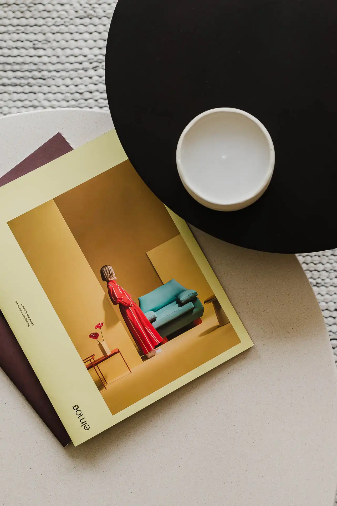 Top-down view of an open design magazine resting on a round two-tone table next to a decorative two-tone ceramic bowl.