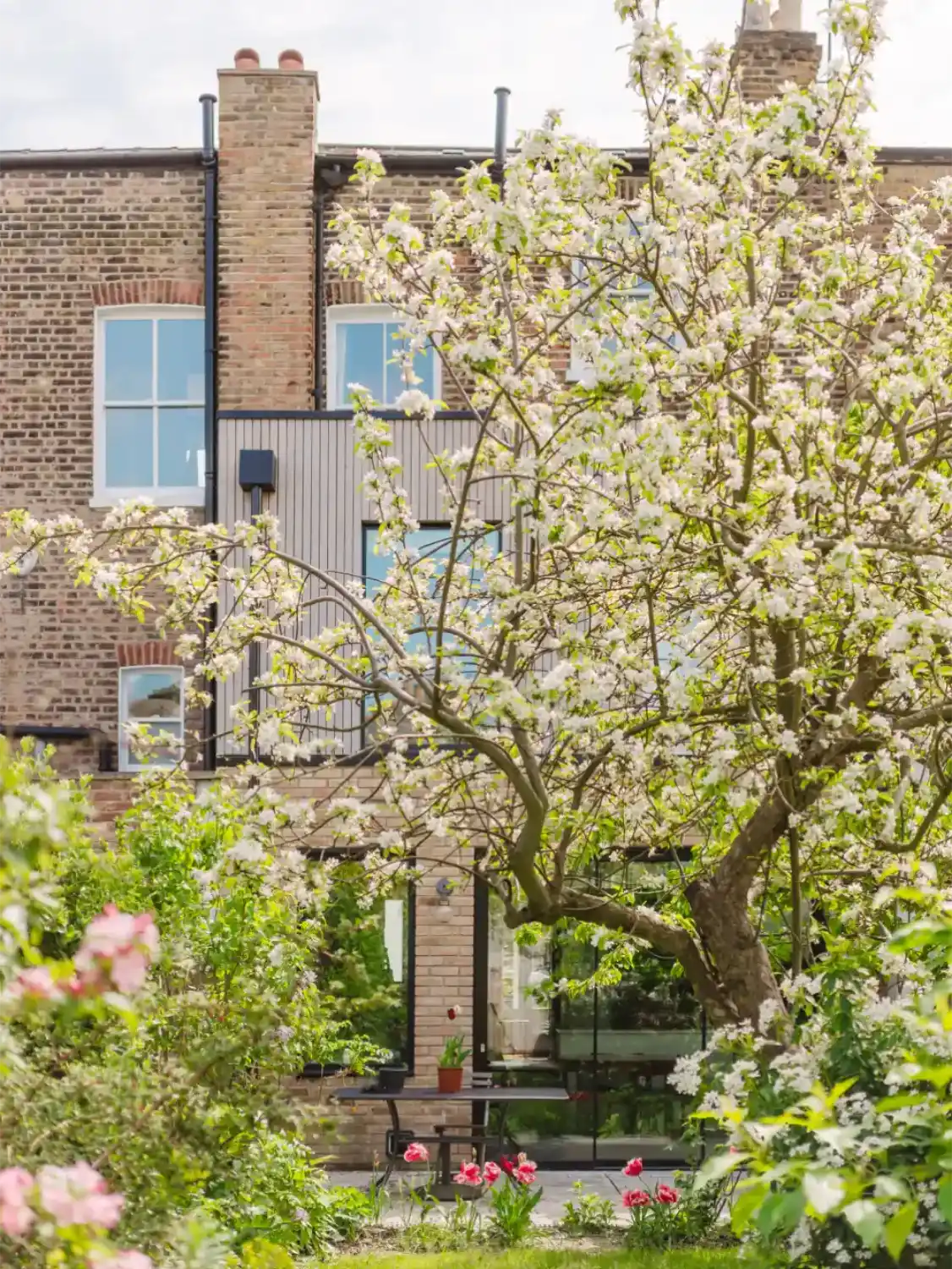 Rear exterior view of a modern brick house extension featuring a timber-clad upper level and glass patio doors, seen through a flowering blossom tree in a garden