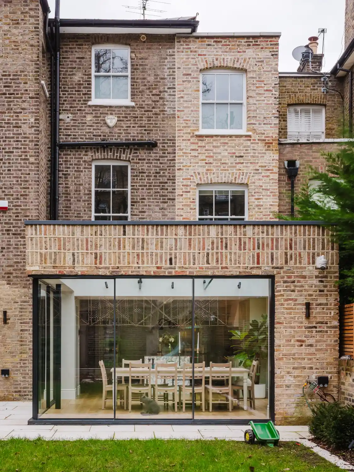 Rear exterior of a brick house featuring a modern, glass-walled ground floor extension that reveals a dining table and chairs inside.